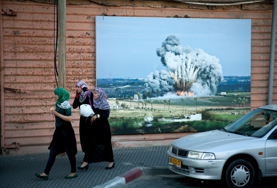 Conservative Palestinian women in traditional dress walk past a brick building displaying a large photograph of an explosion in a rural landscape.