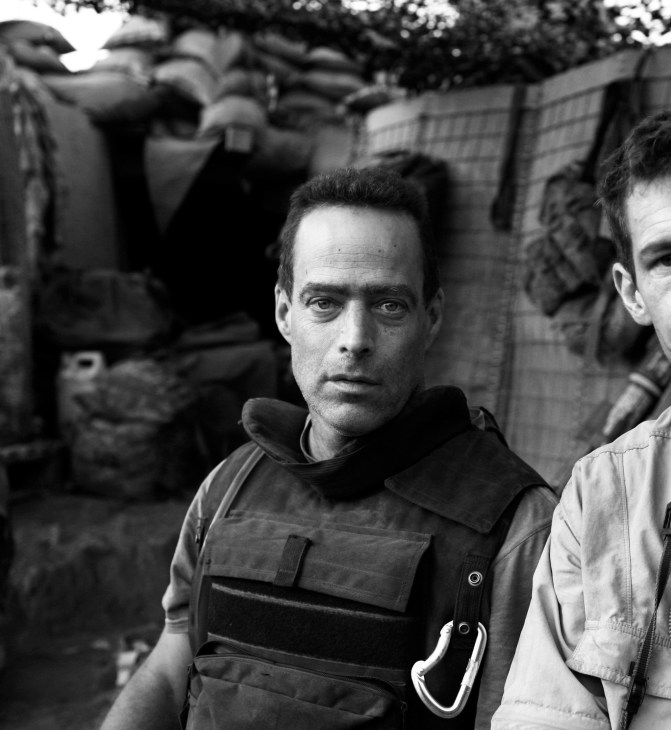 Black and white portrait of Sebastian Junger, a man wearing a dark tactical vest over a black shirt.