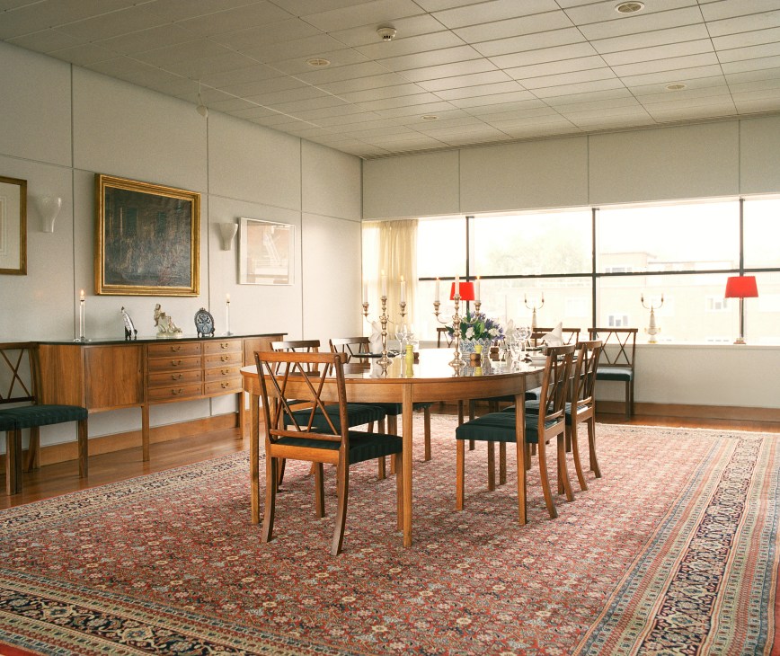 Danish embassy dining room with oval wooden table, mid-century chairs, oriental rug, and expansive windows overlooking London.