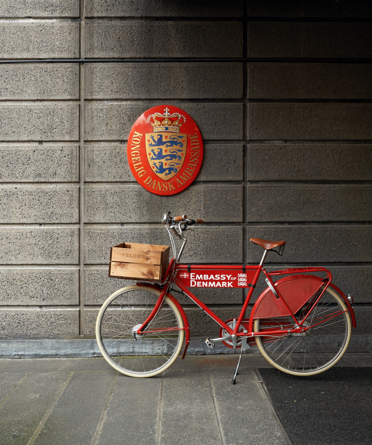Red bicycle with wooden crate and 'Embassy of Denmark' text mounted on brick wall with Danish coat of arms seal above.