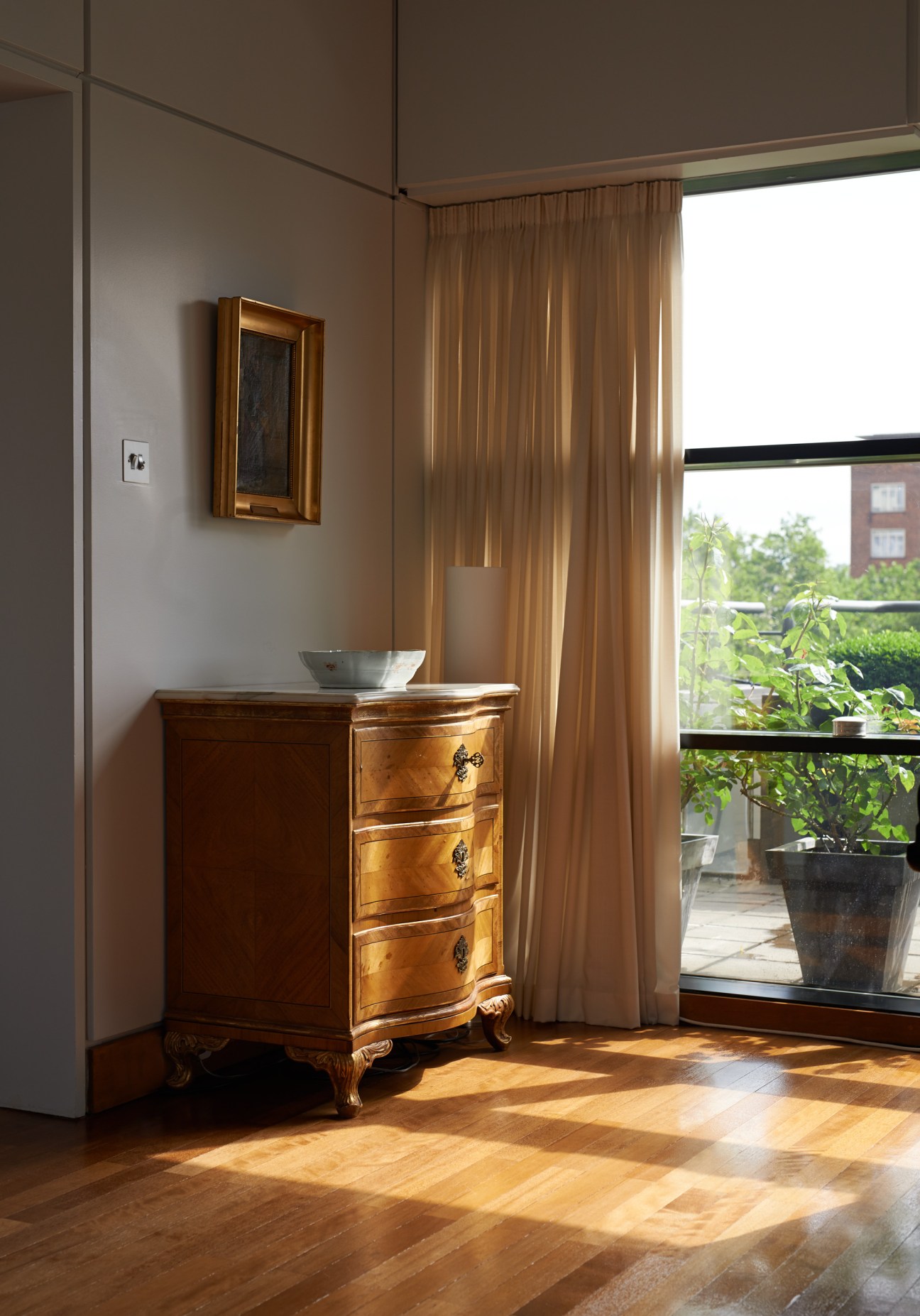 Wooden antique chest of drawers beside framed mirror with golden curtains and window overlooking terrace.