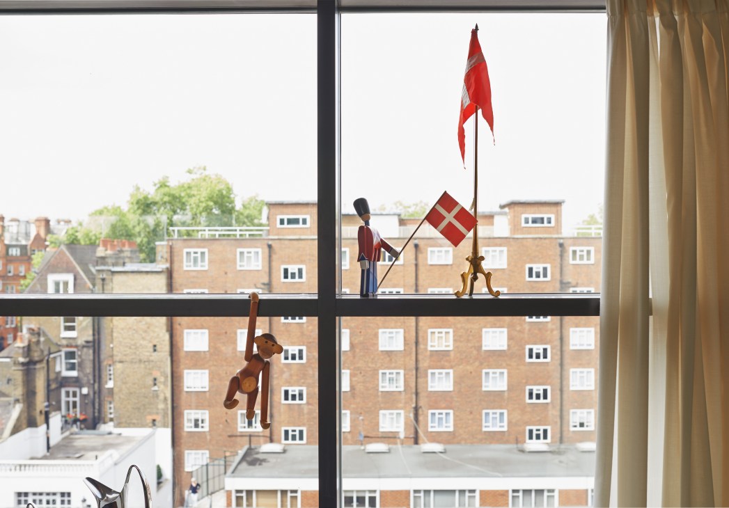 Wooden Boye monkey figurine displayed on a windowsill with Danish flags visible through large windows overlooking London buildings.