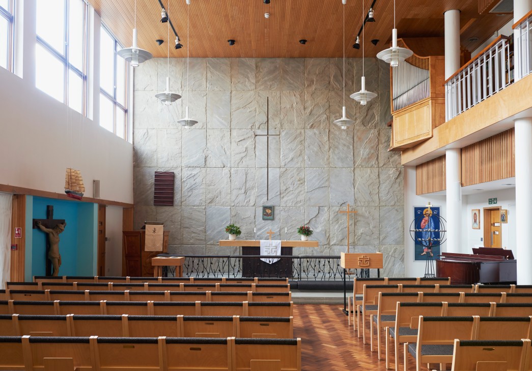 Interior of a modernist church with marble altar wall, wooden pews, and pendant lights hanging from a wood-slat ceiling.