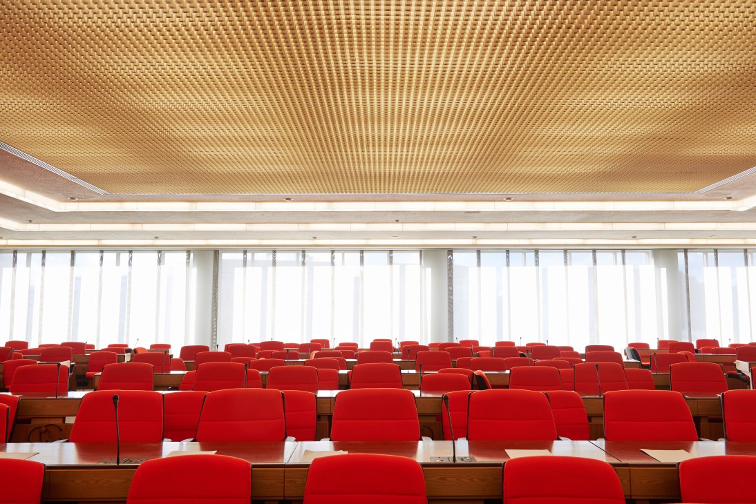 Modern meeting hall with rows of red chairs and a perforated golden ceiling with skylights.