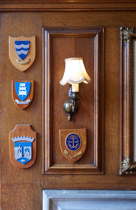 Wooden wall with three maritime ship badges and a brass wall sconce lamp with cream shade.