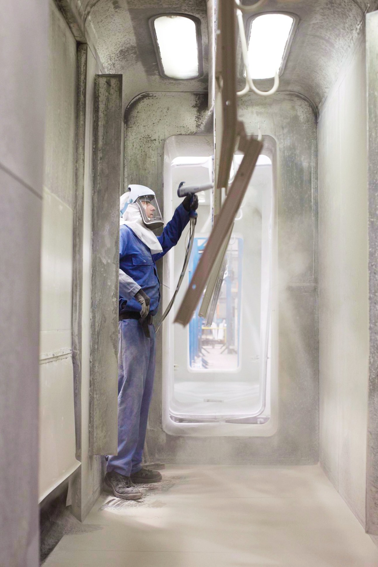 Worker in protective gear spraying polyester powder coating inside a narrow industrial space.