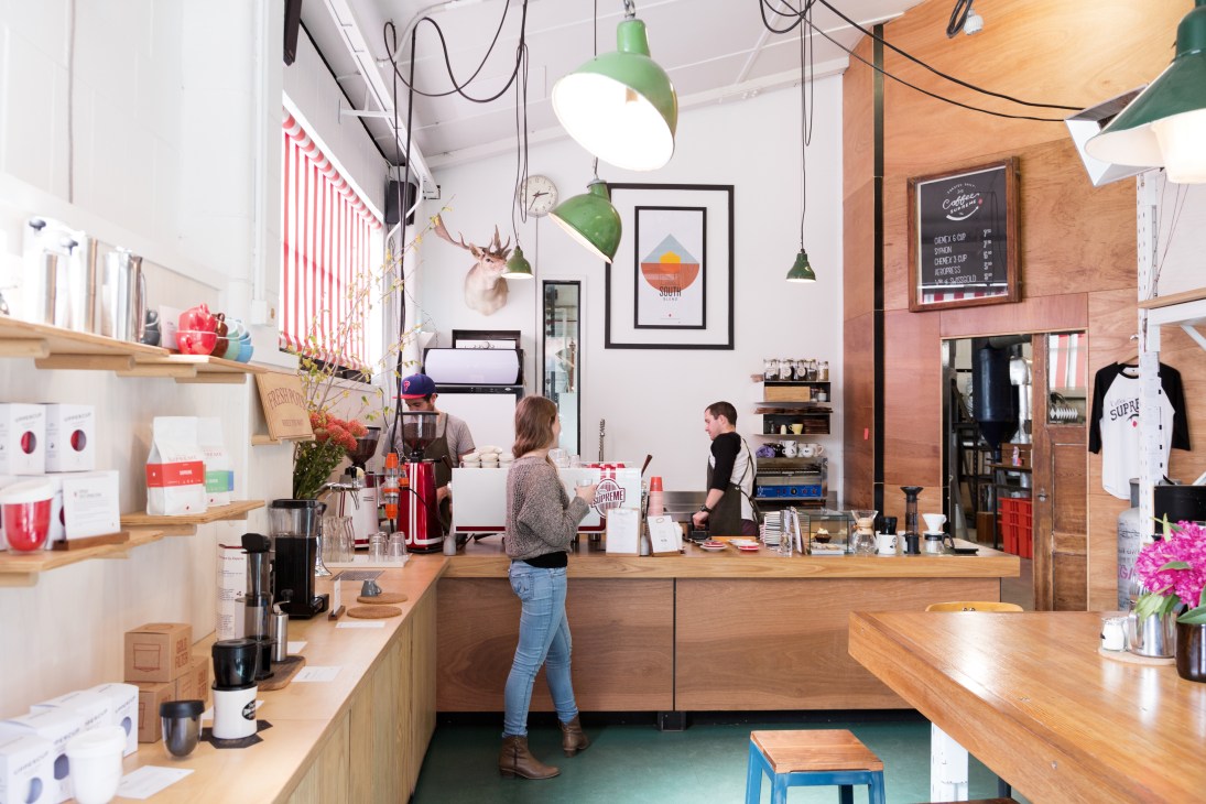 Interior of Abbotsford Club coffee roastery with wooden counter, green pendant lights, and customers ordering.
