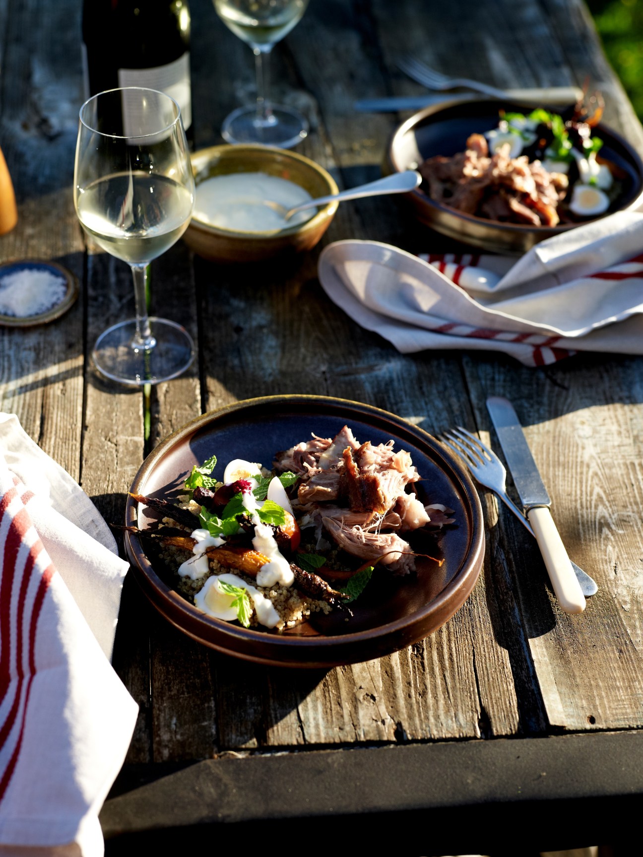 Outdoor dining setup with plated dish of roasted meat and vegetables, white wine glass, and table settings on wooden surface.