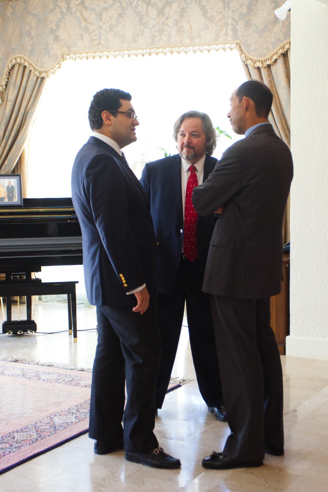 Three men in business suits converse in an elegant room with a piano and draped windows.