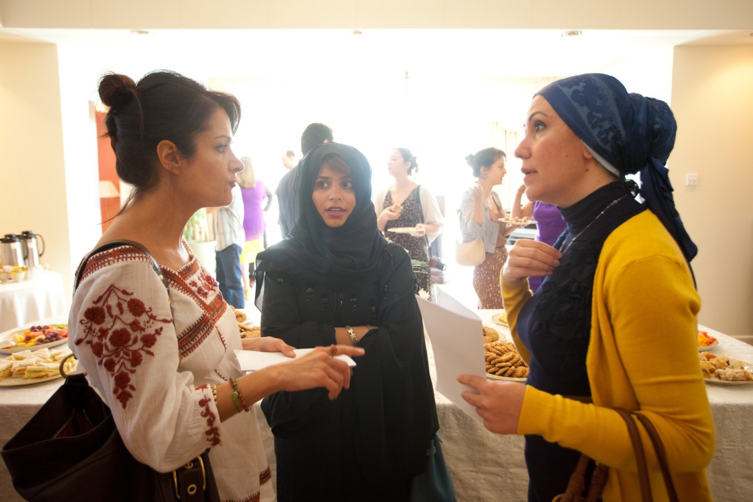 Three women converse at a diplomatic reception with refreshments and other guests visible in the background.