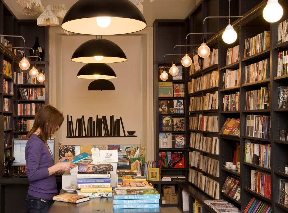Customer browsing books at Lutyens & Rubinstein bookshop counter with shelves and pendant lighting.