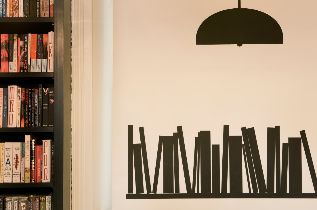 Bookshelf wall decoration with stacked books illustration and hanging pendant lamp at Lutyens & Rubinstein.