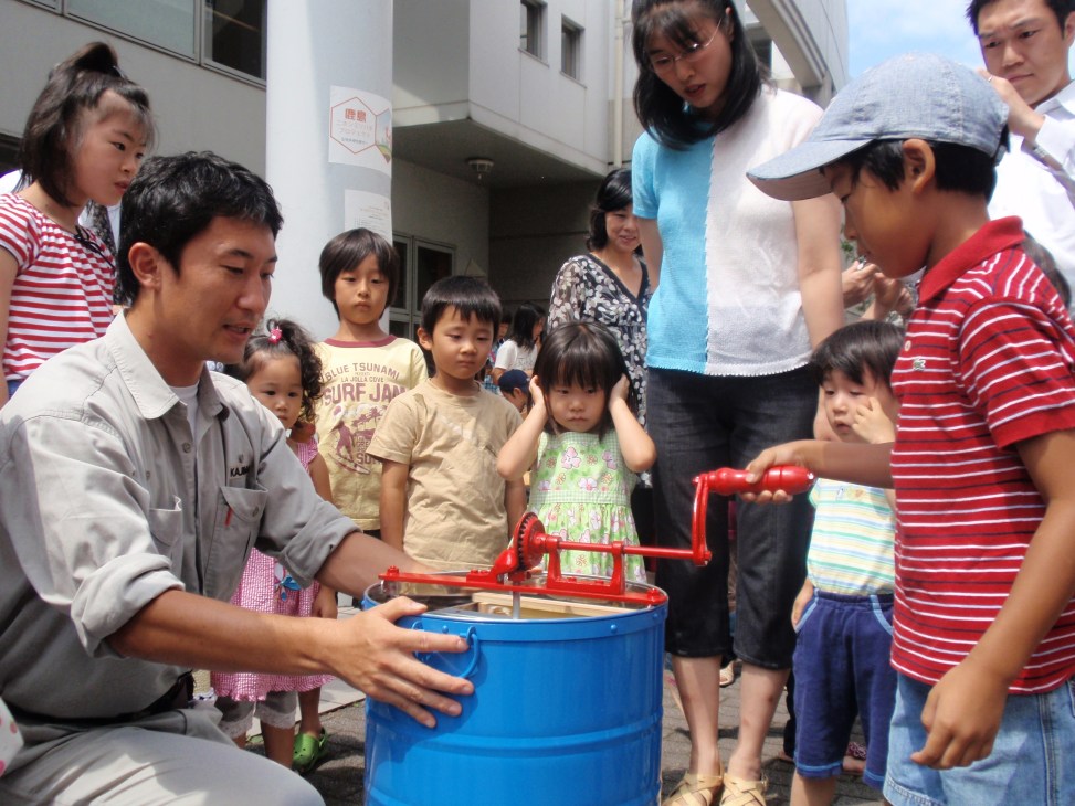 School children gather around an adult demonstrating honey extraction from a blue barrel with a red hand-crank press.