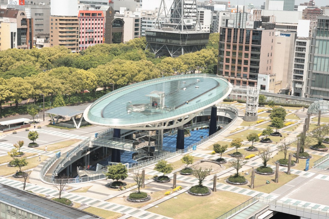 Aerial view of Oasis 21 park in downtown Nagoya featuring a modern oval structure with turquoise water feature surrounded by green landscaping and trees.