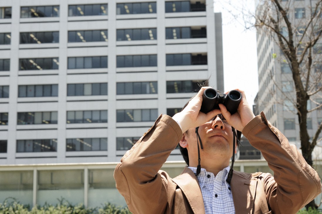 Yoriyuki Yamada uses binoculars to observe bees in trees, with modern office buildings behind him.