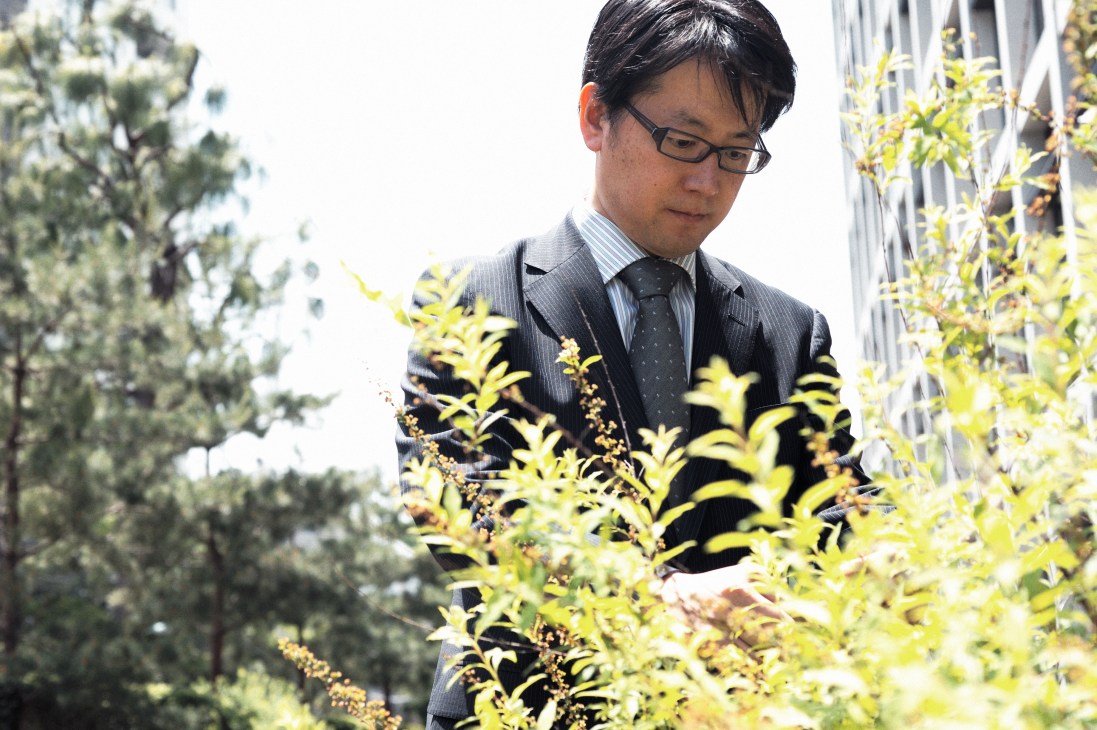 Yoriyuki Yamada in business attire examining flowering plants on a rooftop among green landscaping.