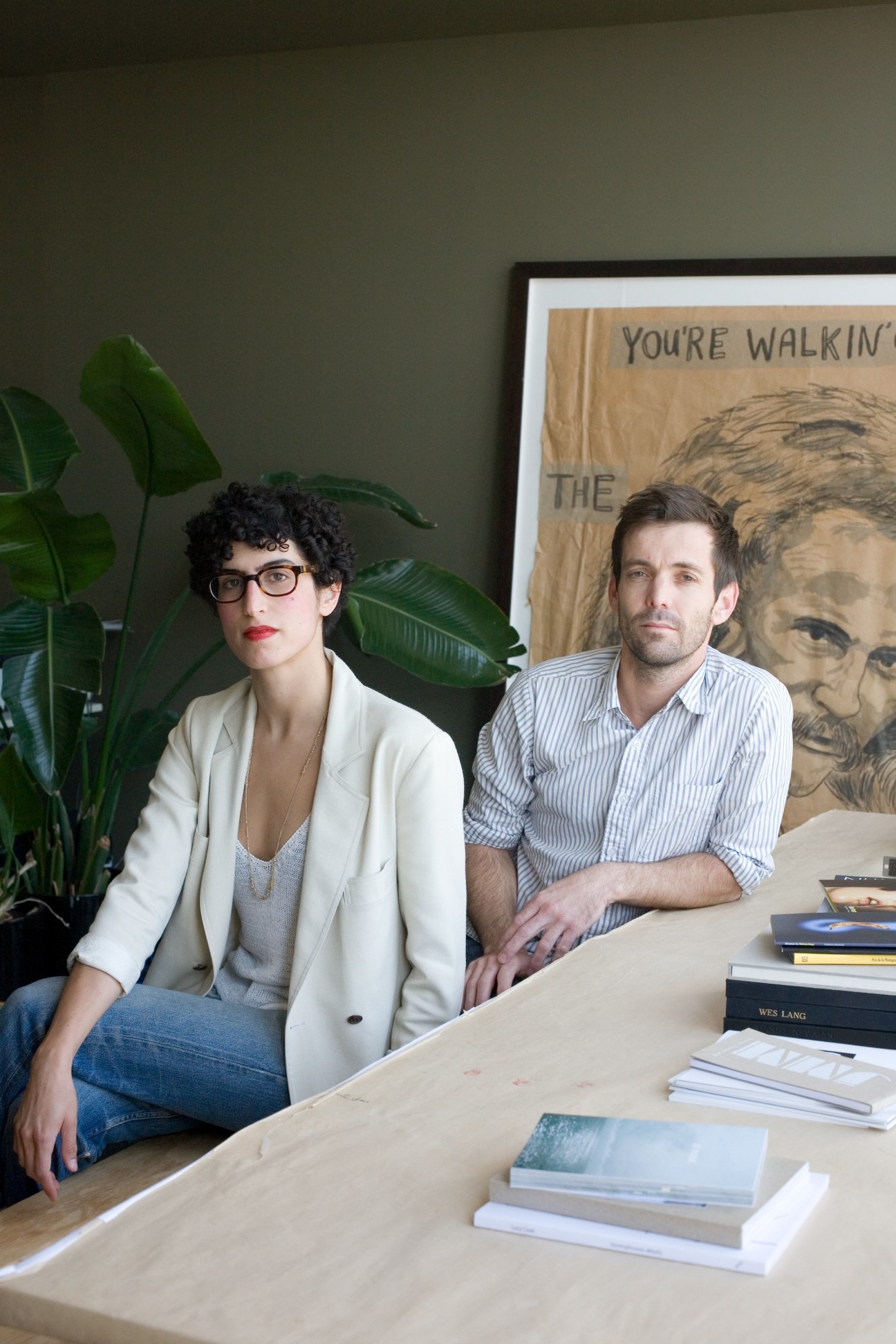 Nicole Katz and Kane Austin sit at a wooden desk surrounded by books in their Los Angeles gallery.