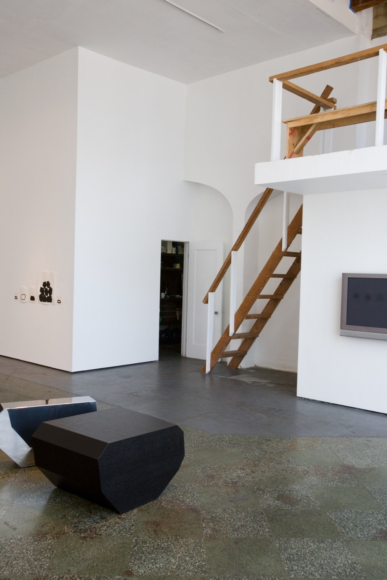 Modern gallery interior with white walls, wooden staircase, and minimalist black sculptural seating.