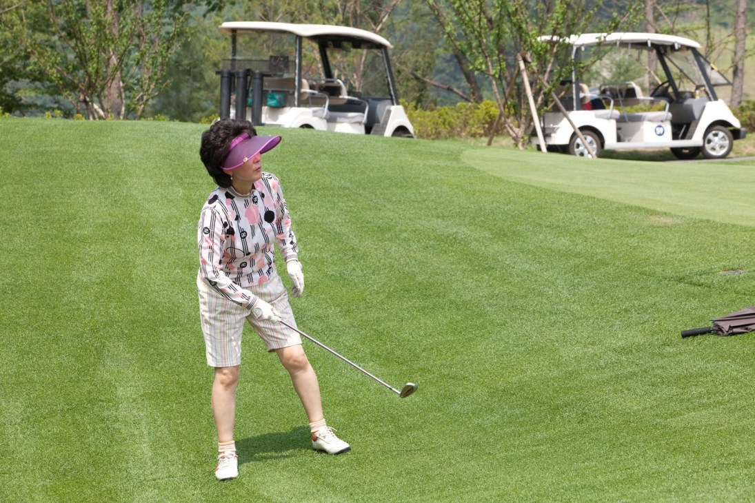 A woman in pink and white patterned golf attire swings on a manicured green fairway with golf carts and trees in the background.