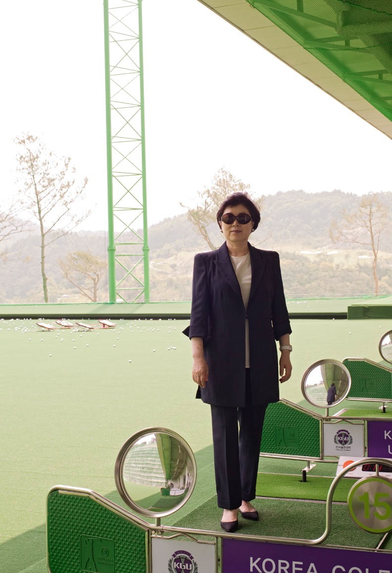 Woman in dark blazer and sunglasses standing at Korea Golf University driving range with mountains in background.