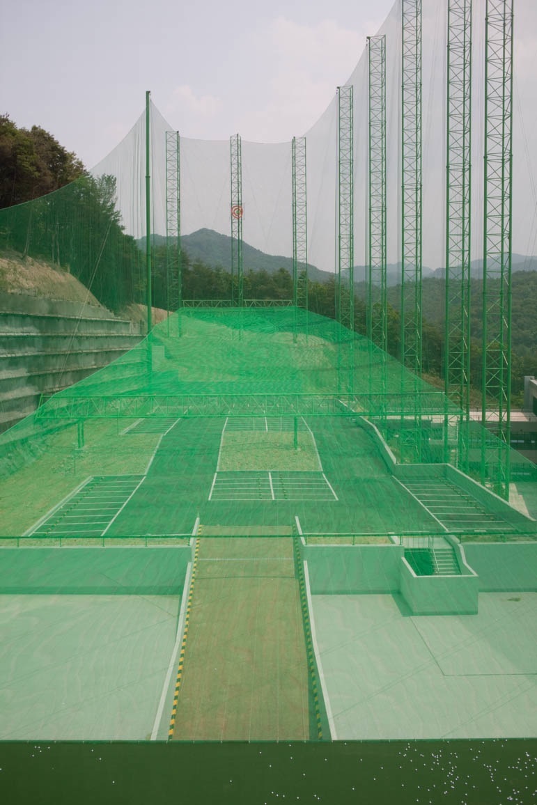 Multi-level golf driving range with green netting and metal framework structures overlooking mountains.