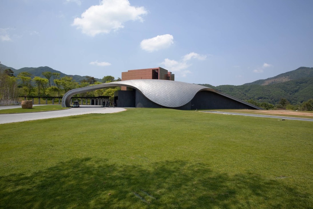 Modern golf clubhouse with curved white roof and black walls set on manicured green lawn with forested mountains in background.
