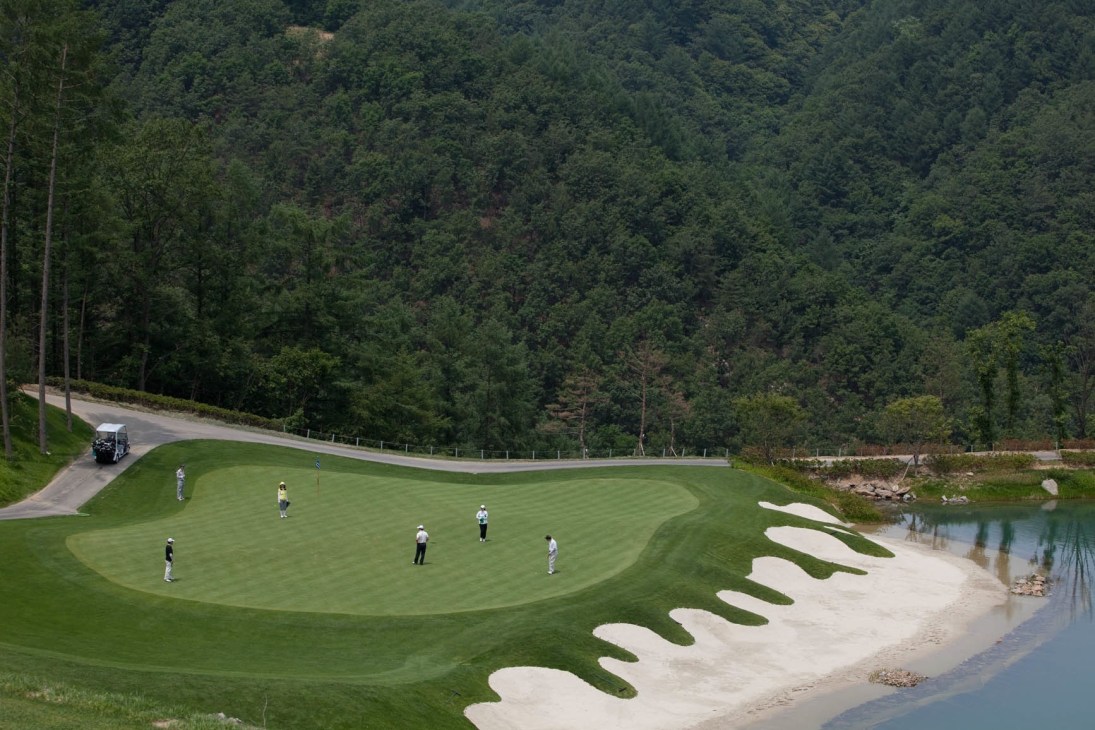 Golfers play on a manicured green course with white sand bunkers beside a stream, forested mountains rising behind.