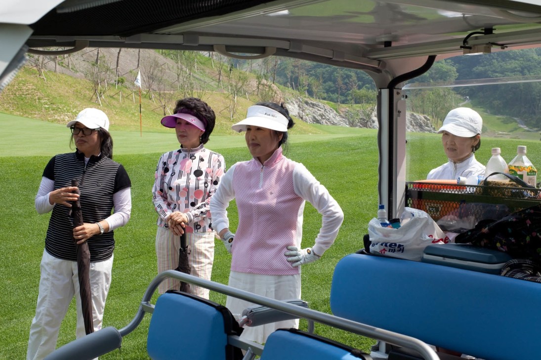 Four women in colorful golf attire stand by a golf buggy on a course with mountains in the background.