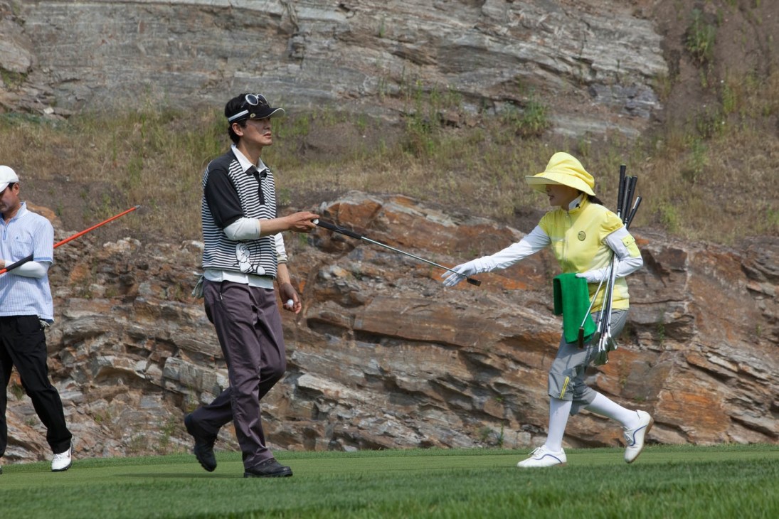 A female caddy in a yellow visor and white sleeves hands a golf club to a male golfer in striped shirt and cap on a course with rocky cliff backdrop.