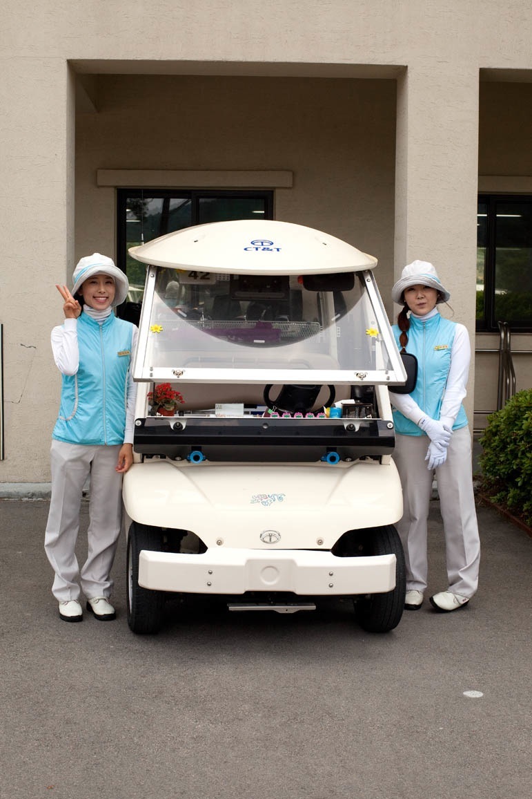 Two female caddies in light blue and white uniforms stand beside a white golf cart at ChungWoo Golf Club.