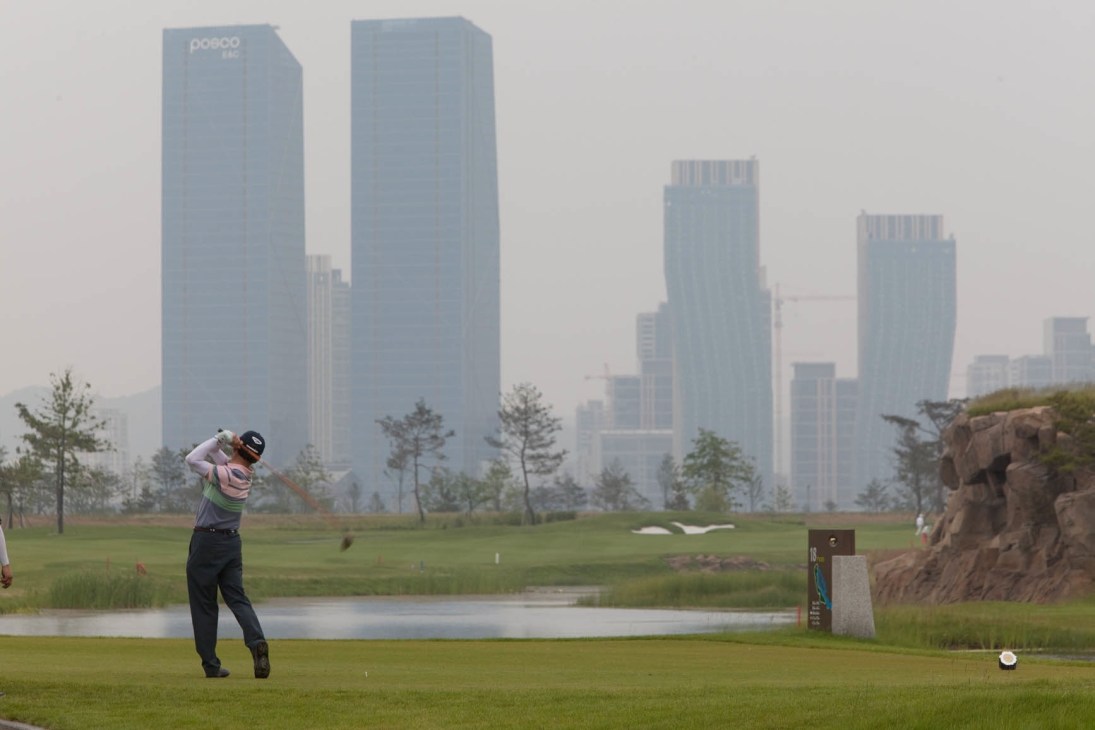 Golfer mid-swing at Jack Nicklaus Golf Club Korea with modern skyscrapers in the background.