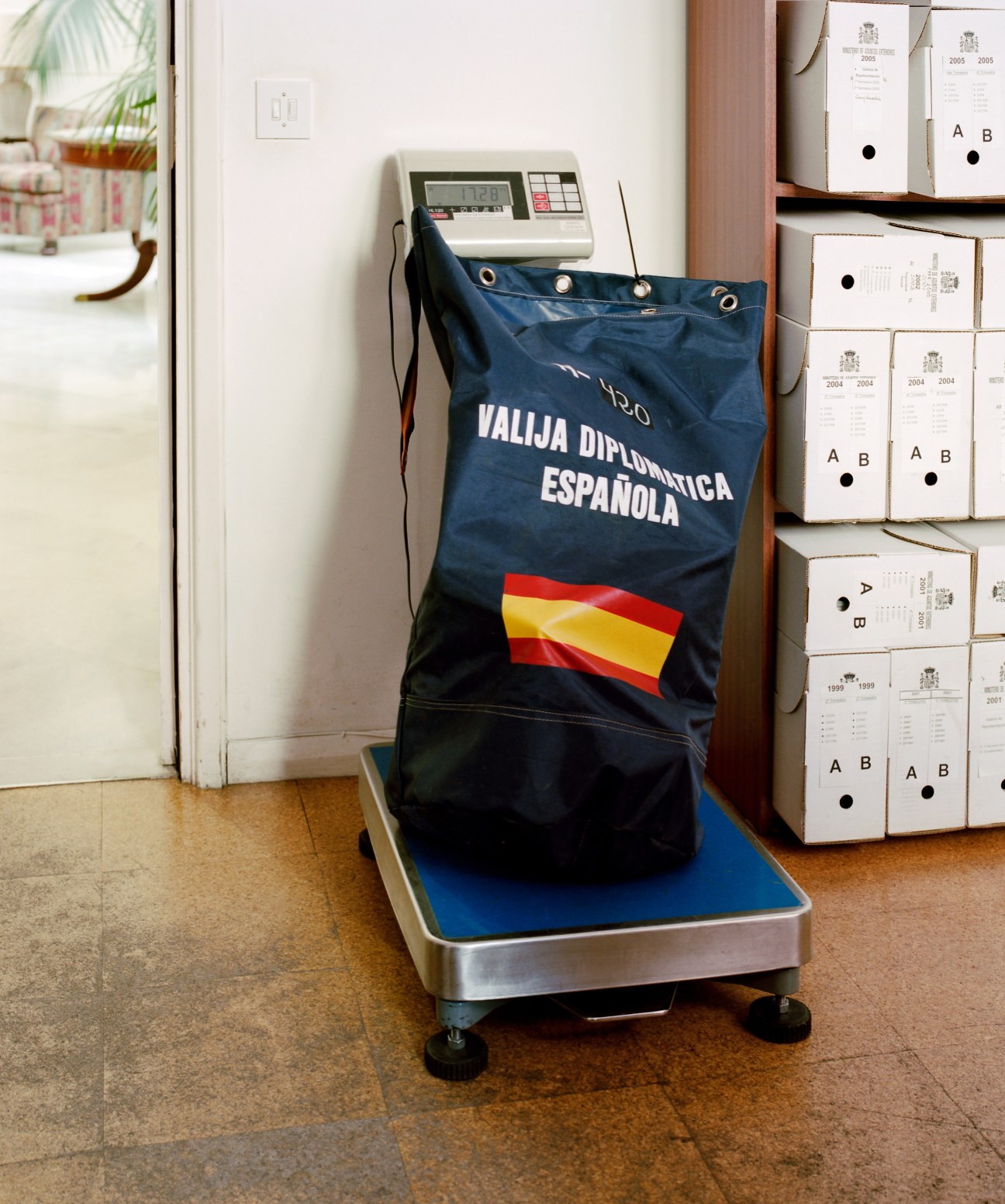 Spanish diplomatic pouch labeled 'VALIJA DIPLOMATICA ESPAÑOLA' being weighed on a scale at an embassy.