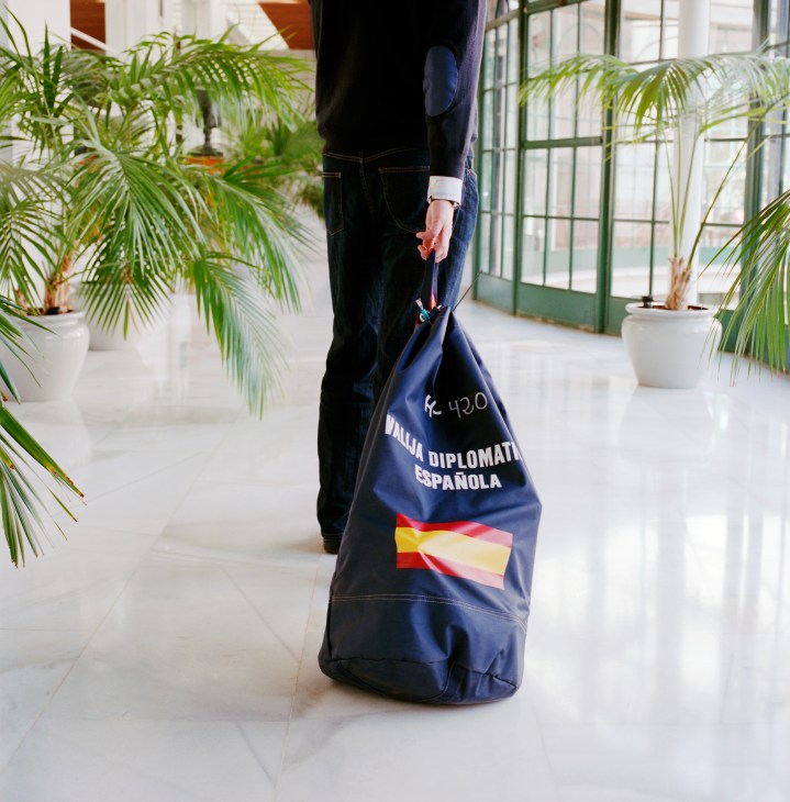 A person carries a blue Spanish diplomatic pouch marked 'Pouch Diplomática Española' with the Spanish flag through a bright corridor with potted plants and green-framed windows.