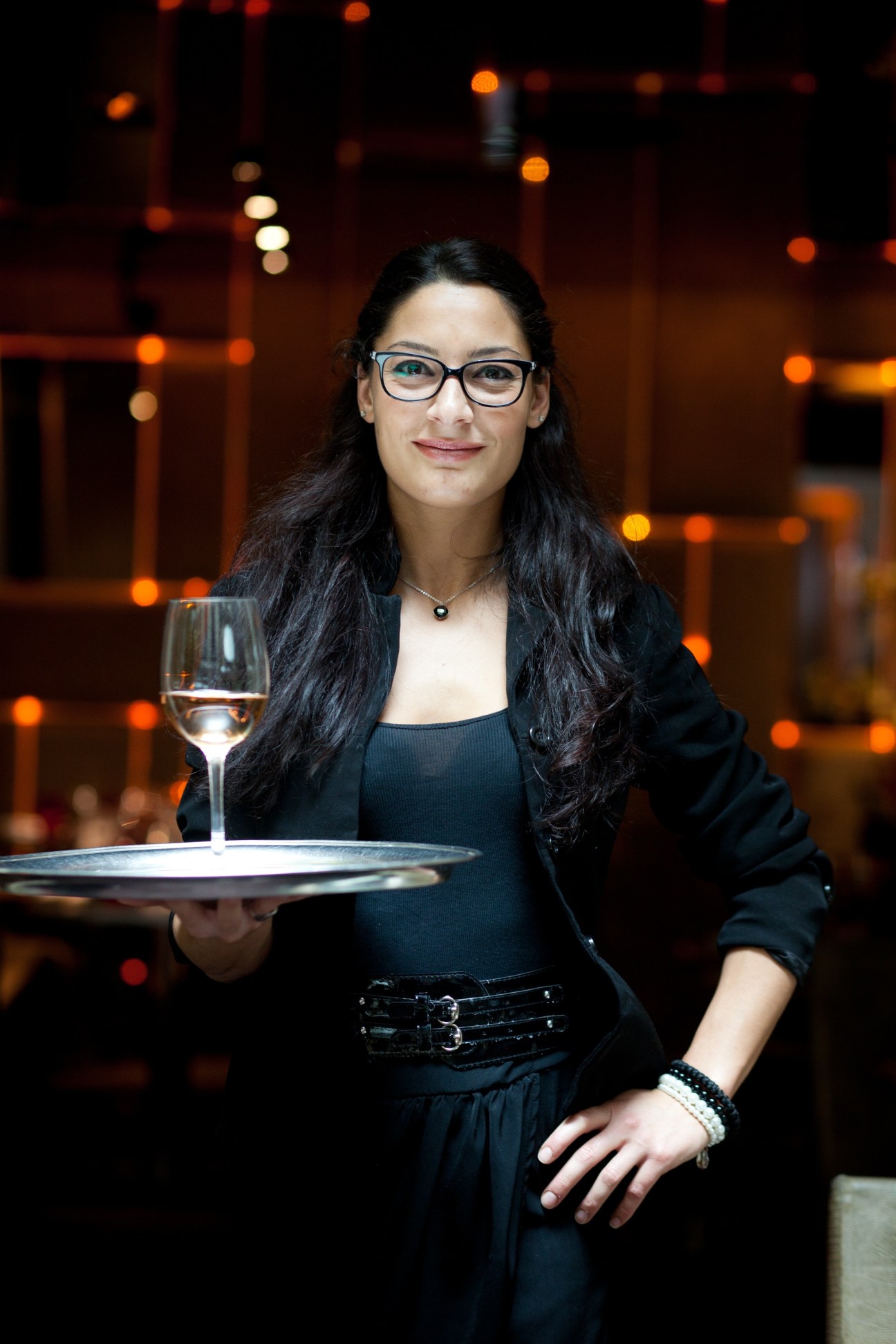 A server in black attire holds a tray with a glass of rosé wine in an upscale restaurant setting.