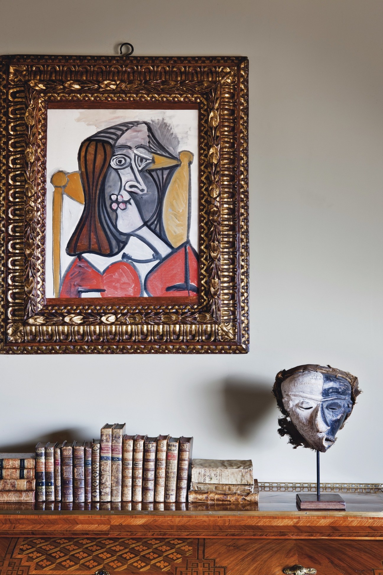 Framed Picasso-style cubist portrait painting displayed on wall above antique books and sculptural head on wooden shelf.