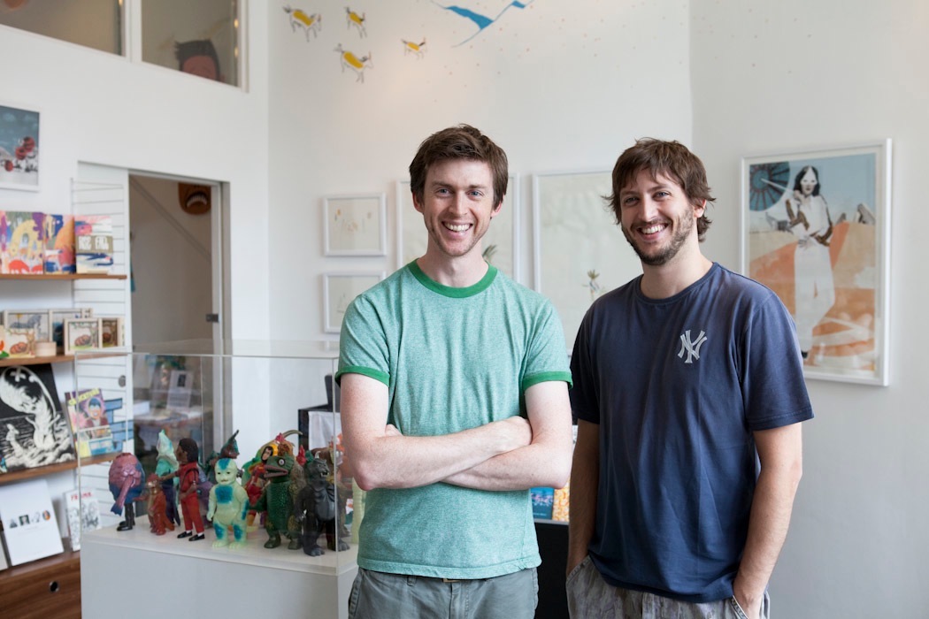 Nobrow founders Sam Arthur and Alex Spiro standing in their studio with colorful action figures and artwork displayed behind them.