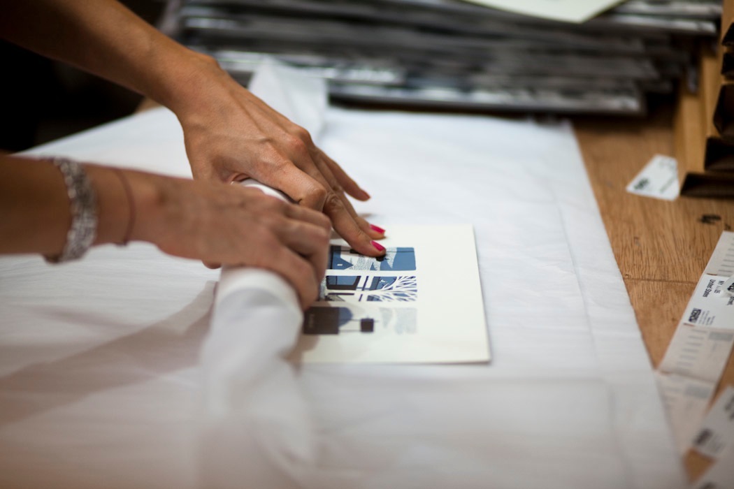 Hands wrapping printed materials with blue illustration design on white paper at a wooden table.
