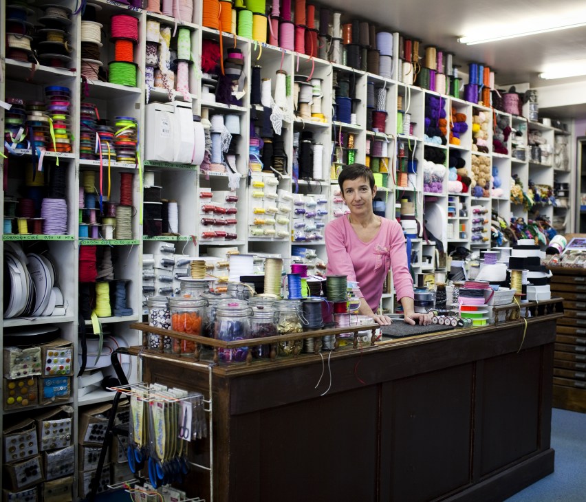 Sophie Andrieux stands at the counter of La Mercerie de Charonne, a haberdashery shop with colorful thread spools organized on white shelves behind her.