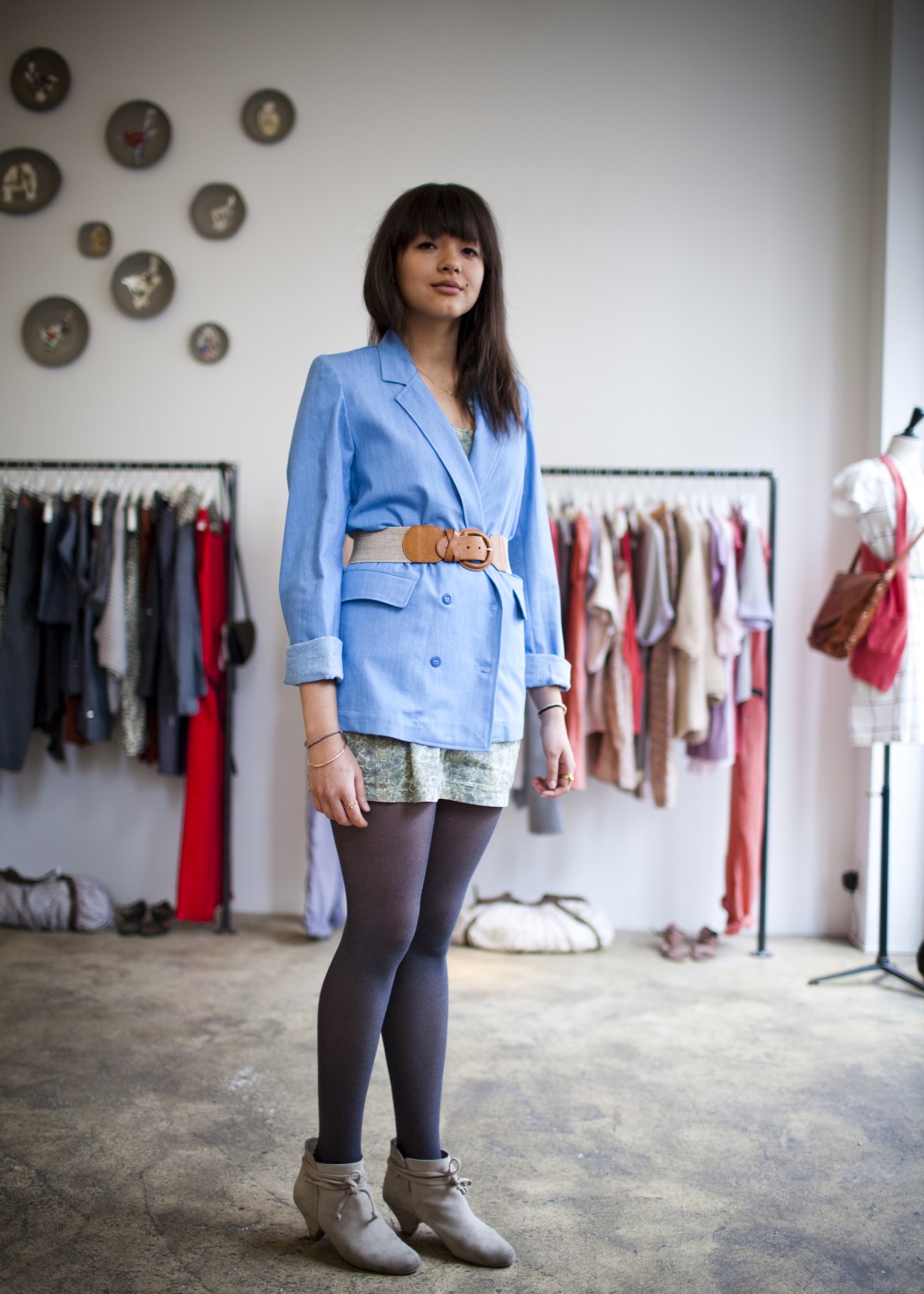 Shop assistant wearing a blue blazer and tan belt stands in a Sessùn boutique with clothing racks in background.