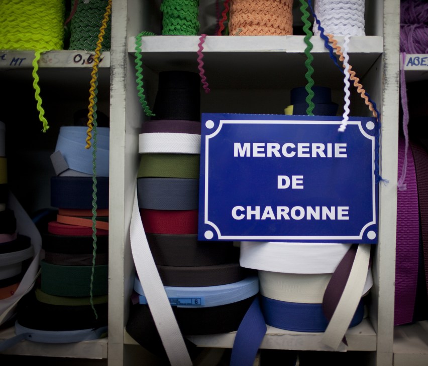 Blue enamel sign reading 'MERCERIE DE CHARONNE' displayed among colorful ribbons and trims in a haberdashery shop window.