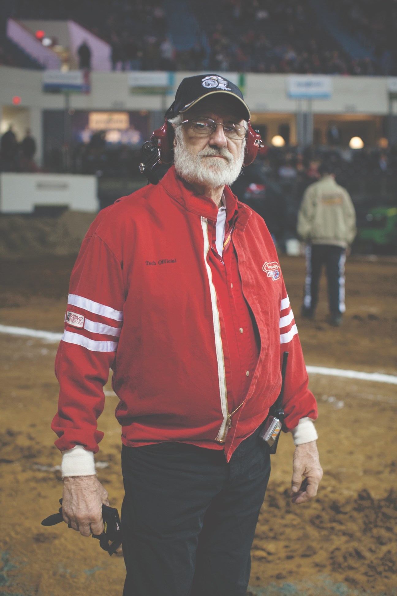 An elderly man in a red official jacket and cap stands in a dirt arena at a tractor pull event.
