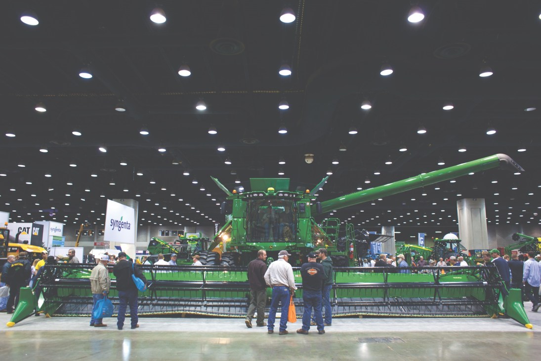 Farmers gather around a large green John Deere S-series combine harvester at an indoor exhibition.