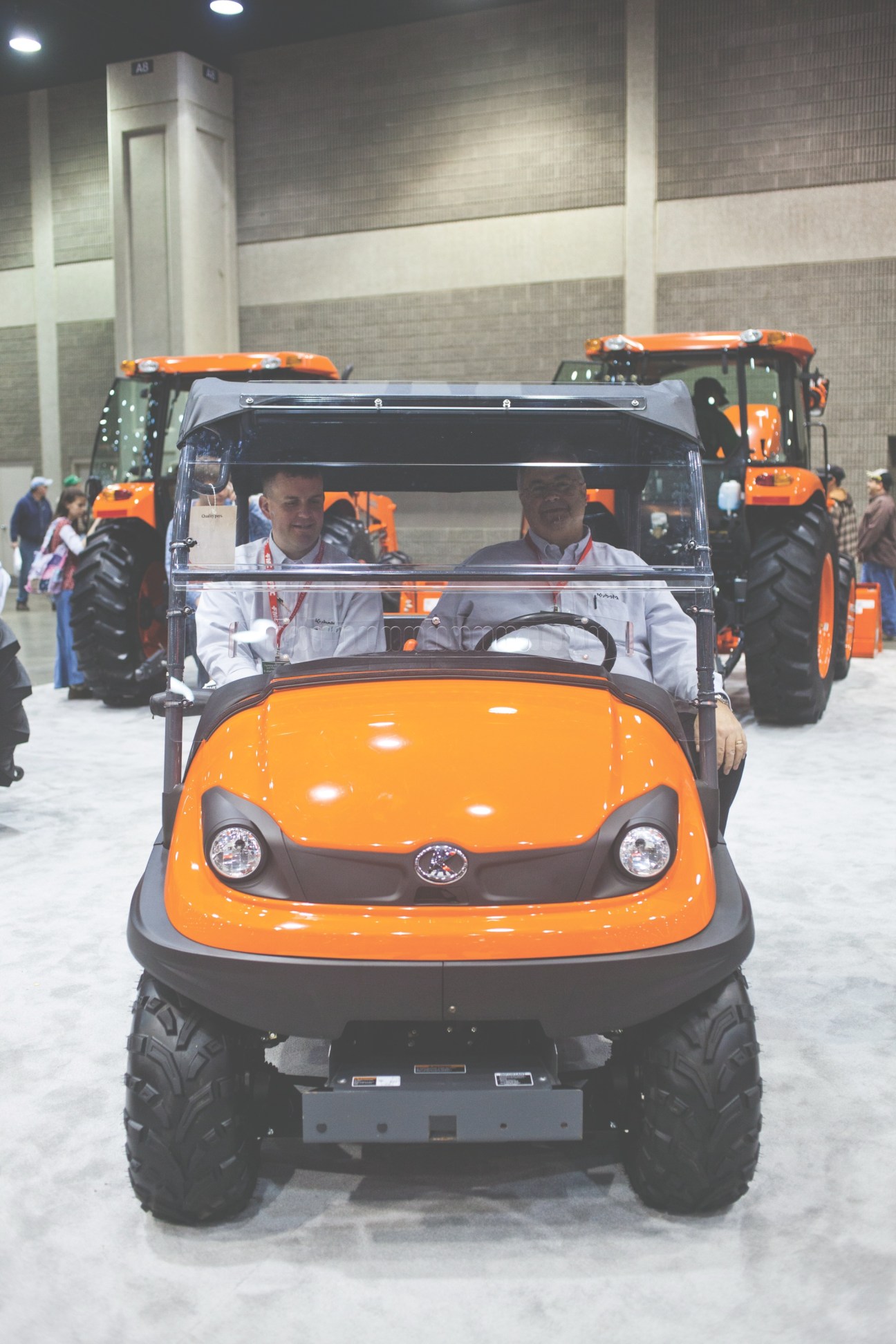Two Kubota salesmen sit in an orange RTV utility vehicle at an agricultural exhibition.