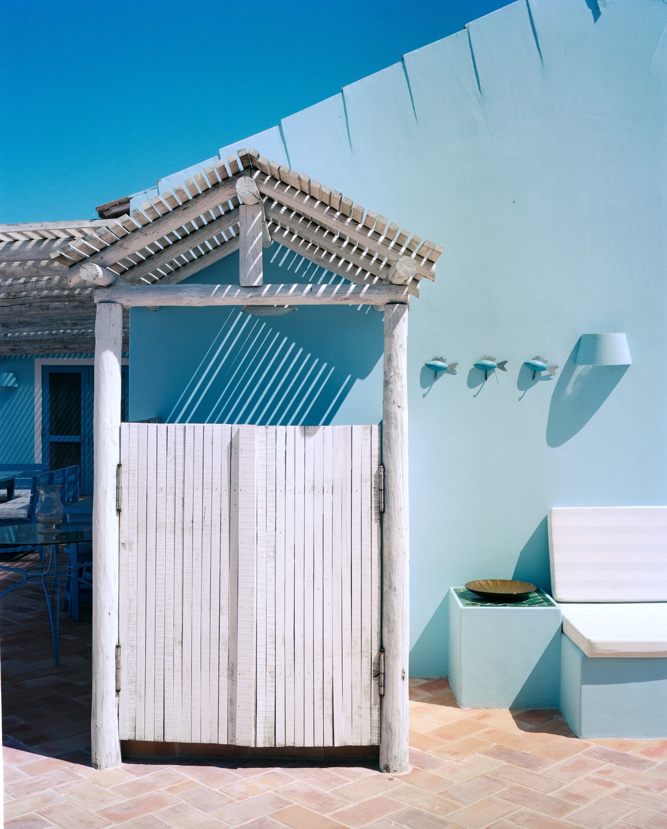 A white wooden changing hut with blue door and pergola roof against a turquoise wall, with decorative bird sculptures and white shelving visible.