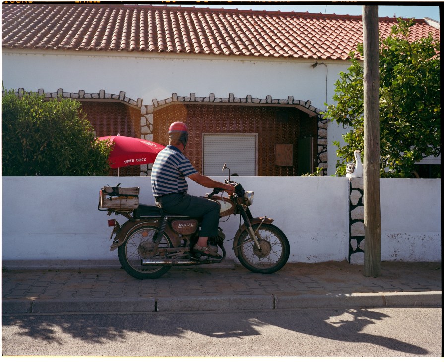 Man riding a scooter with a red surfboard past a white-washed house in Comporta.