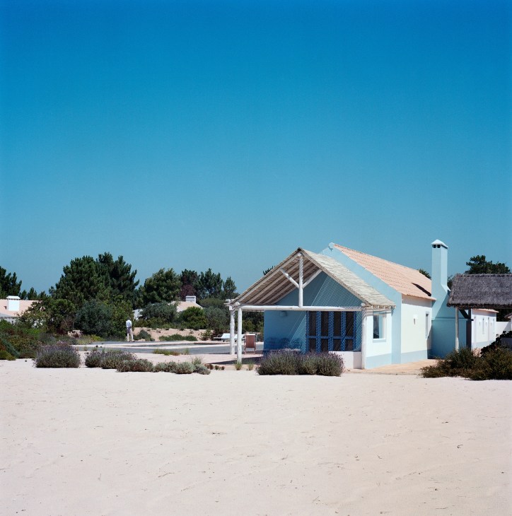 Modern beach house with white walls and blue shutters on sand, featuring reed roof section and pine trees beyond.