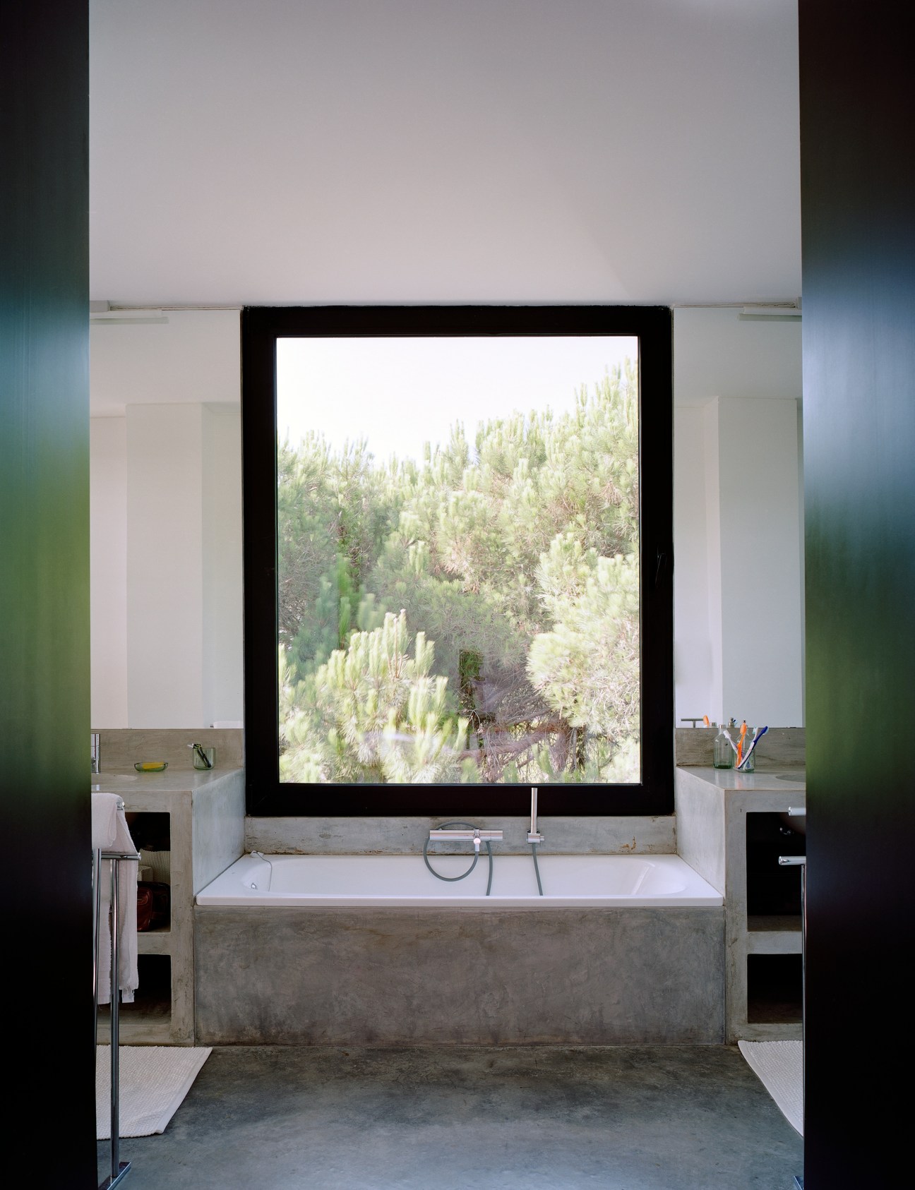 Modern bathroom with concrete bathtub and large black-framed window overlooking pine trees.