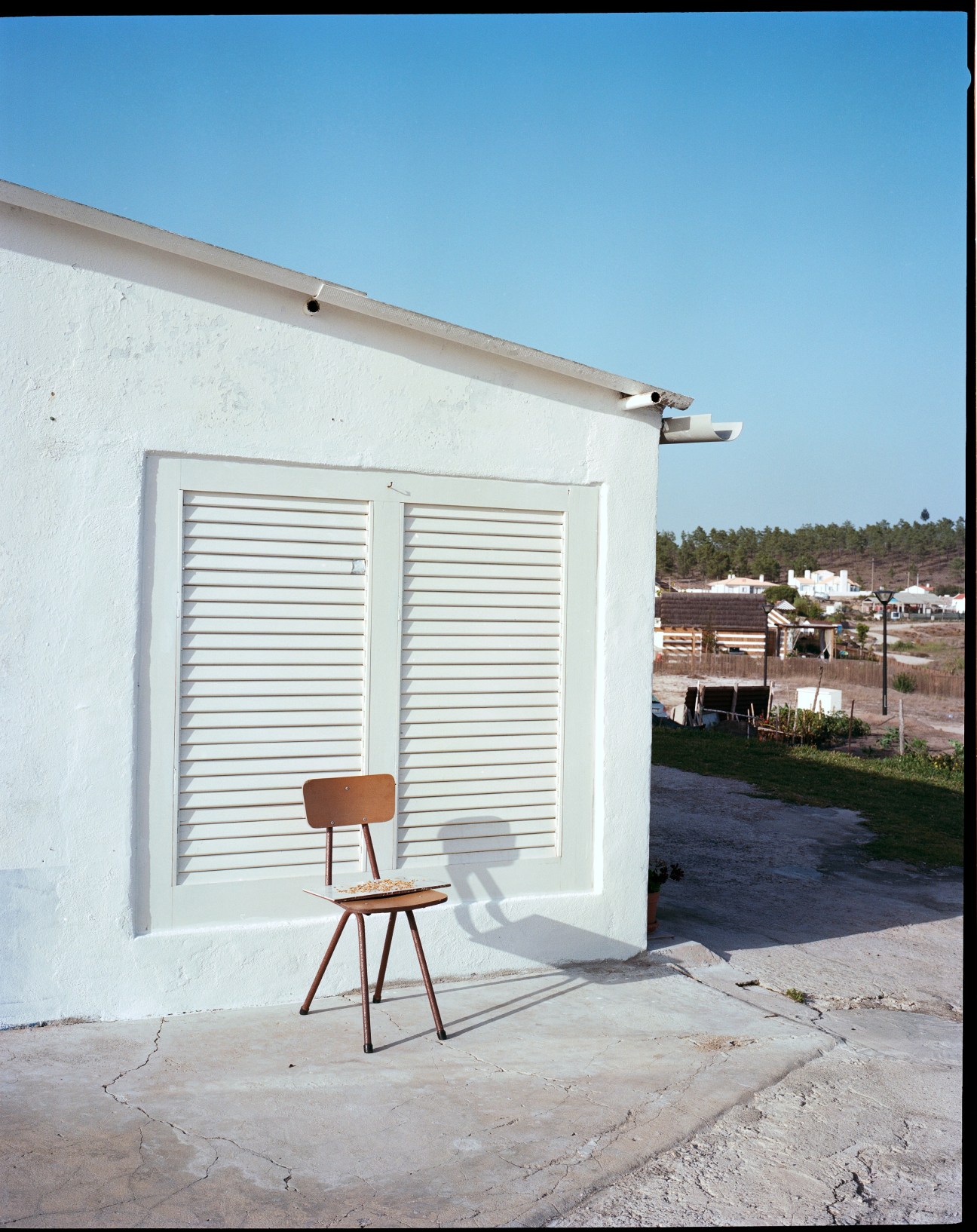 A wooden chair sits in front of a white-walled structure with louvered doors under clear blue sky.