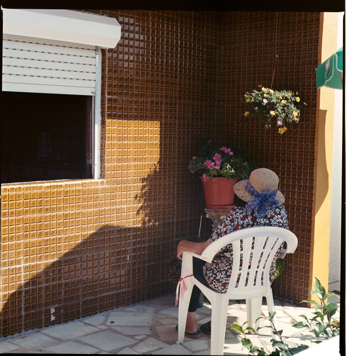 Person in straw hat sitting in white plastic chair in shaded brick courtyard with potted flowers.