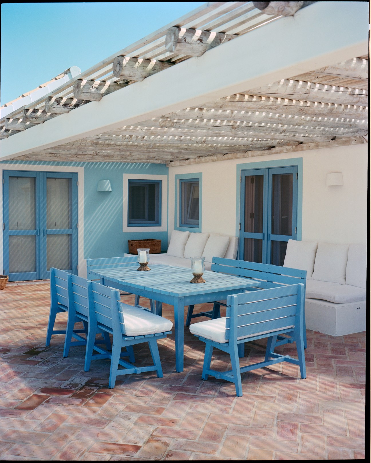 A coastal veranda with blue wooden furniture, white cushions, and blue-trimmed doors overlooking pine trees.